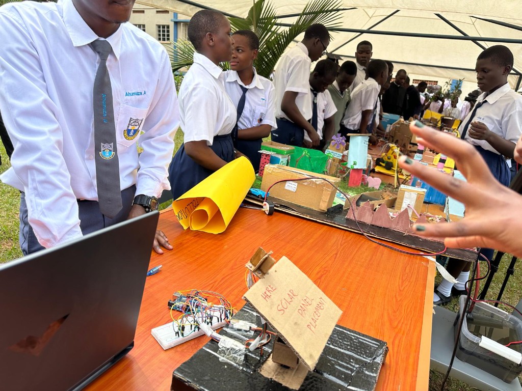 Students at an outdoor science fair presenting projects including solar panel placement wiring, microcontrollers, and models on a display table